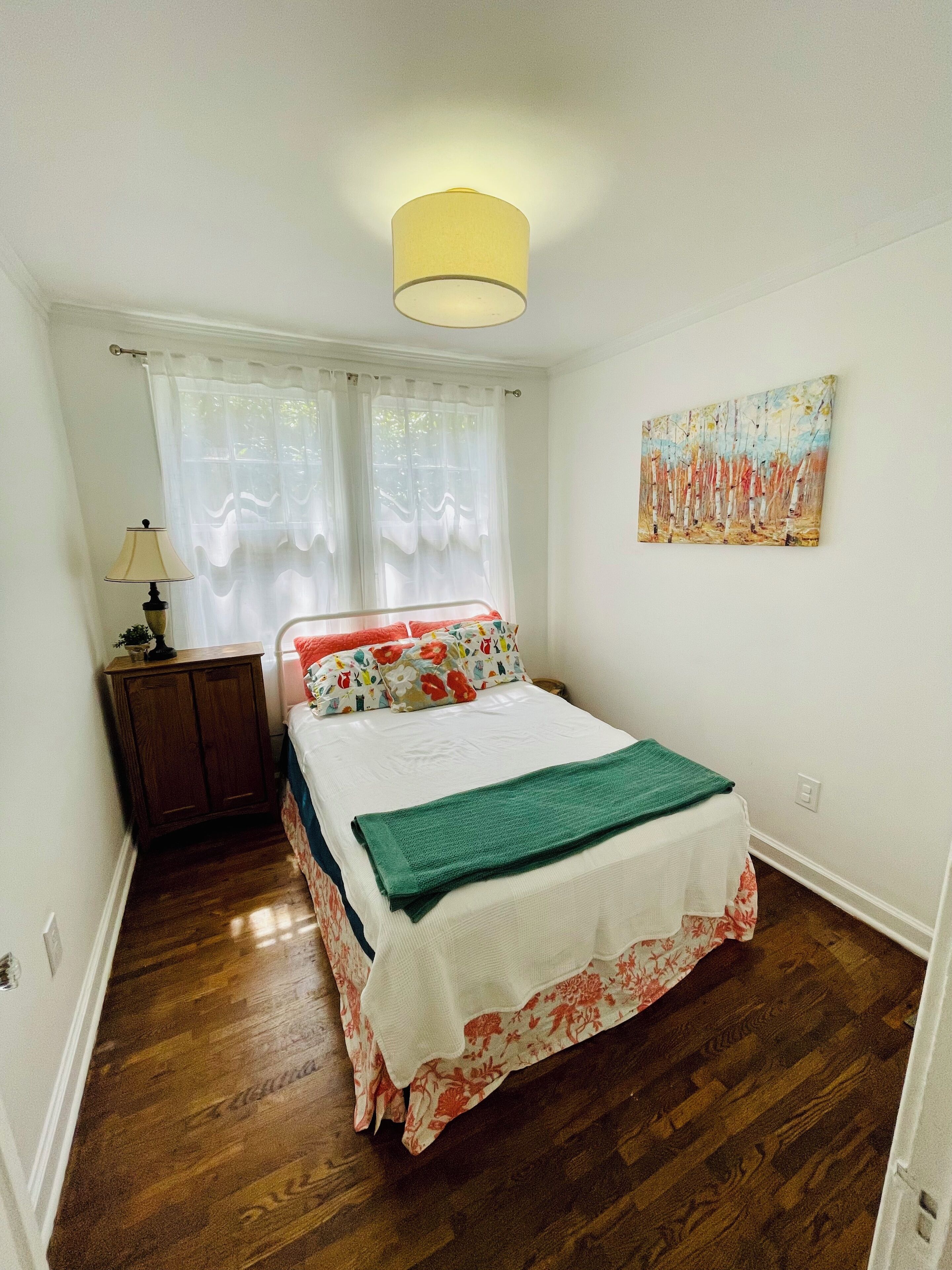 A bedroom with a bed, a nightstand, a lamp, a painting, and a window with curtains.