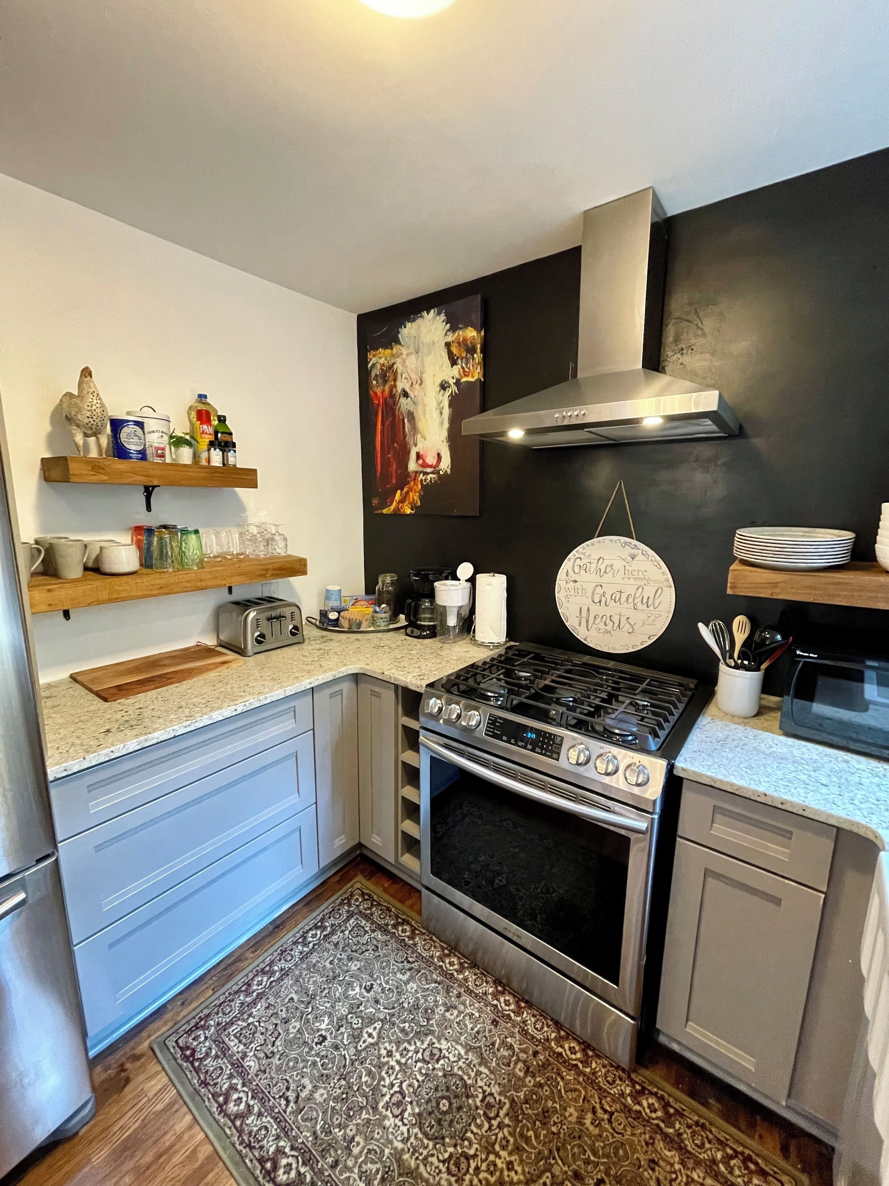A modern kitchen with a gas stove, oven, and microwave. There are wooden shelves with kitchen items, a rug, and a wall-mounted sign.