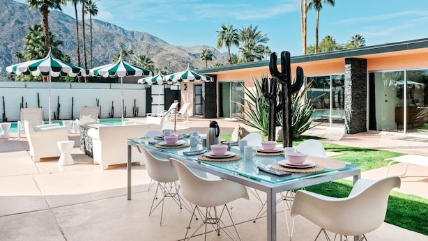 An outdoor dining area with a glass table set for four, surrounded by white chairs, palm trees, and a mountain backdrop.