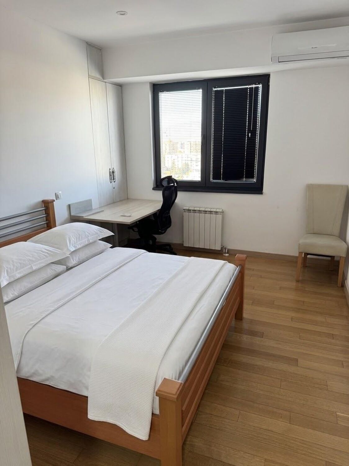 Ein Schlafzimmer mit einem hölzernen Bett, einem Schreibtisch, einem Stuhl und einem Fenster mit Jalousien.