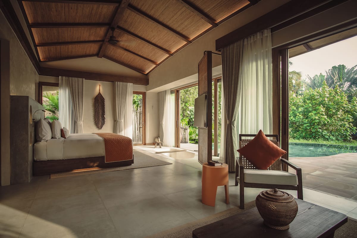 Un dormitorio moderno con cama, una silla, una mesita y vistas a una piscina y vegetación.