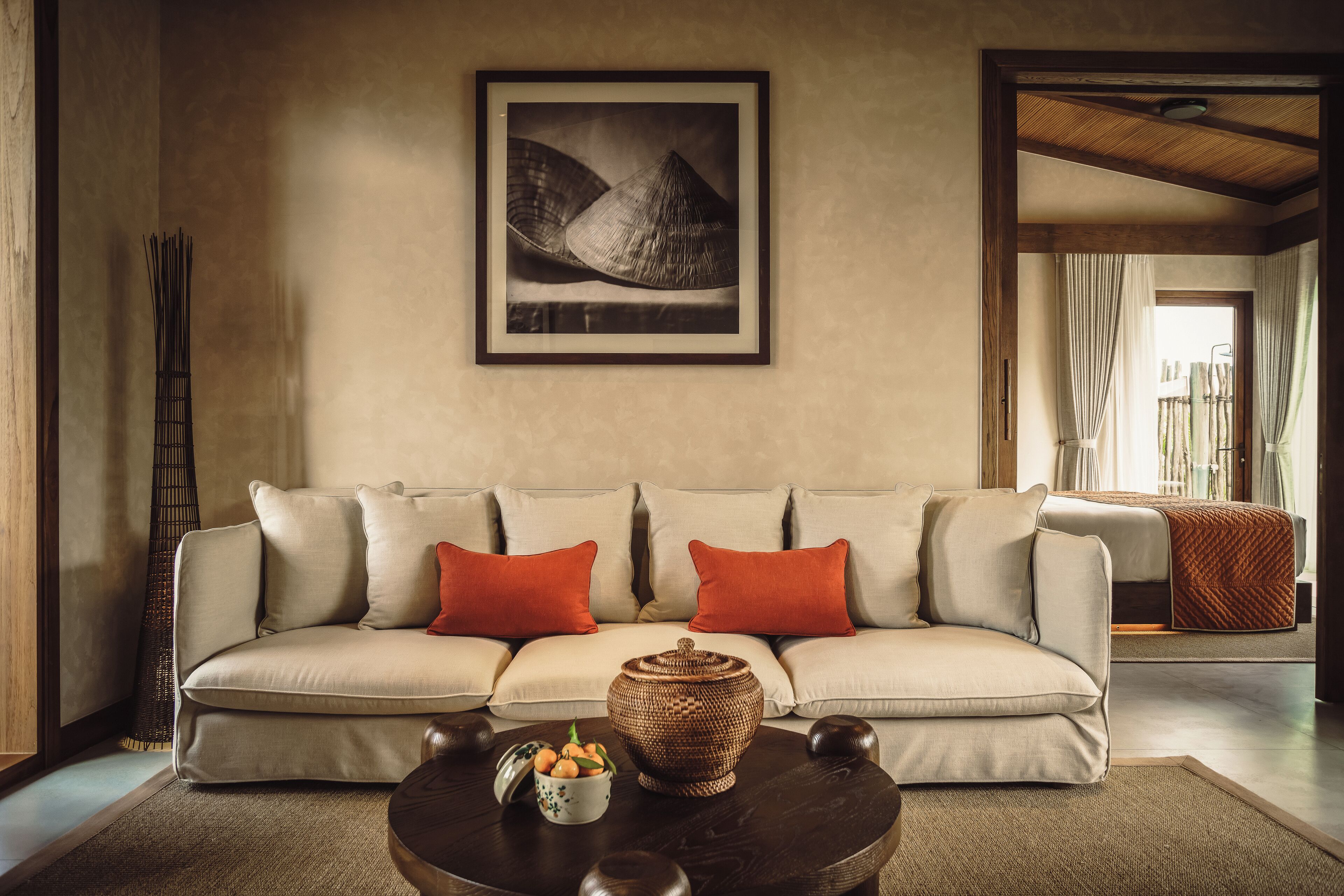 A living room with a beige sofa, two red pillows, a coffee table with a basket and a bowl of fruit, and a framed picture on the wall.