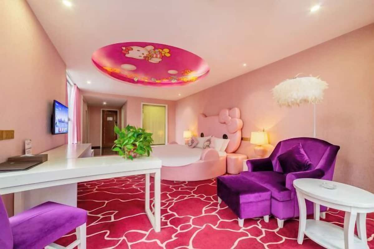 Habitación de hotel con decoración en tonos rosa y morado, cama redonda, escritorio con planta, televisión y adorno en el techo.