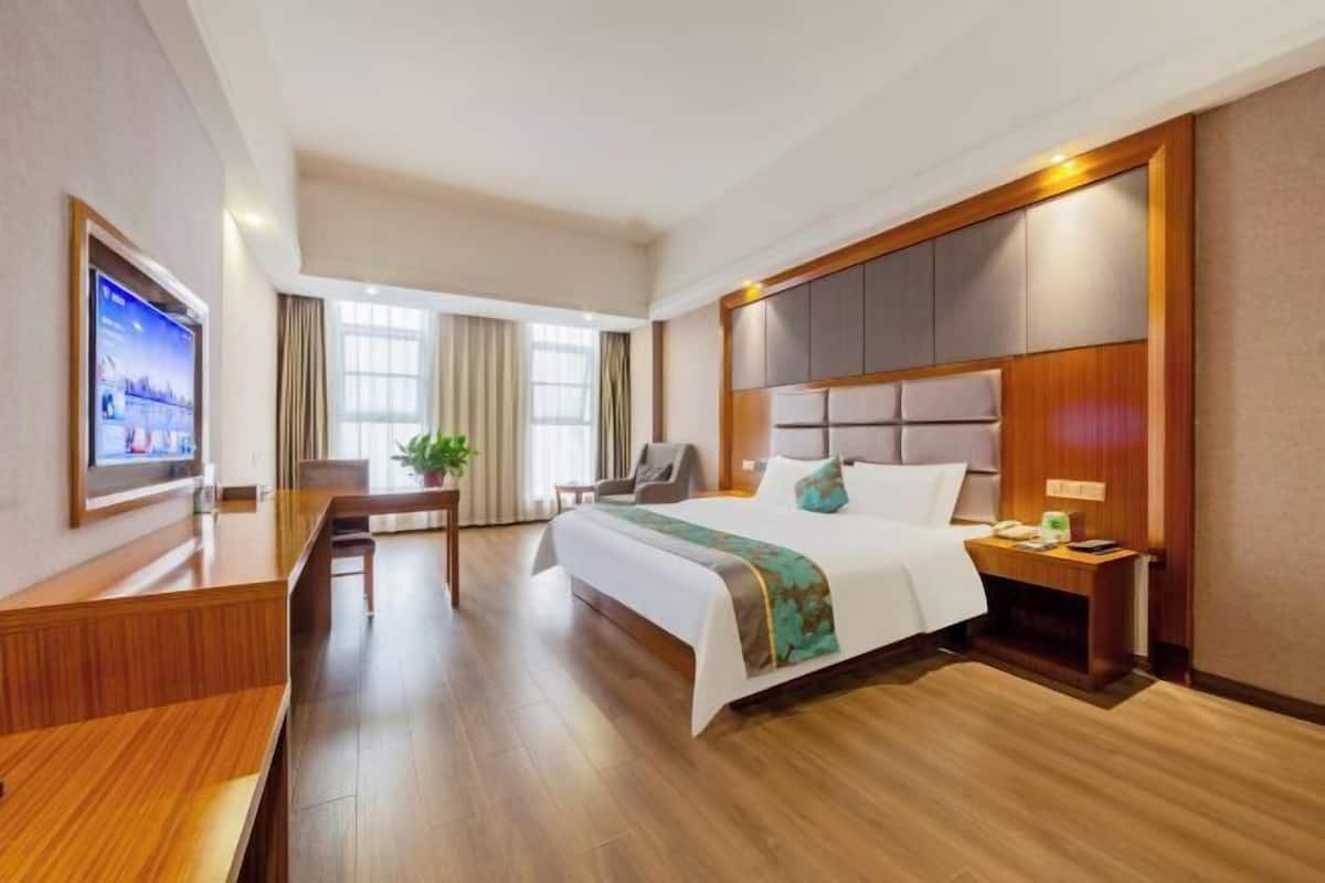 Una habitación de hotel con cama, mesita de noche, televisor, escritorio, silla y ventanas amplias.