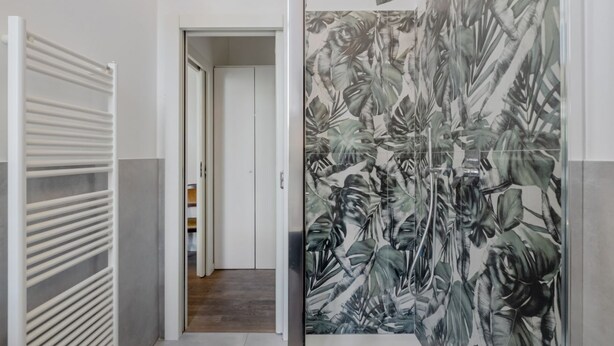 A hallway with a white radiator, a door with a tropical leaf pattern, and a view into a room with a wooden floor. - GuestHost - Modern Apartment 150m from Turro M1 (Milano)