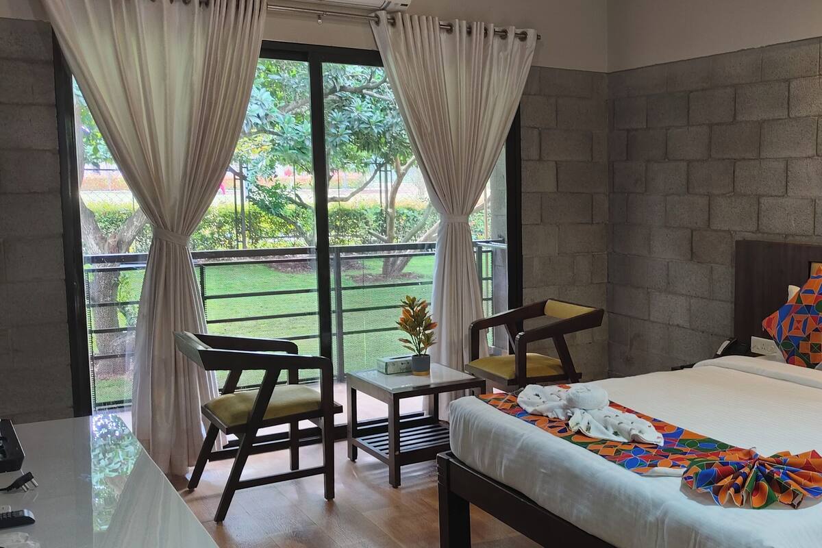 A hotel room with a bed, a desk, chairs, a view of the outdoors, and a painting on the wall.