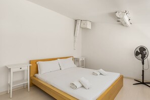 Een enkel bed met witte lakens, een houten hoofdbord, twee nachtkastjes, een wandairconditioner en een ventilator.