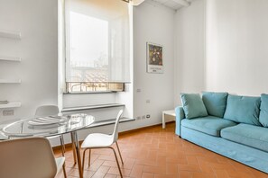 A modern living room with a glass dining table, white chairs, a blue sofa, and a window with a city view. - GuestHost - Charming 80 sqm apartment, for up to 8 people, located on the 4th floor of a building without a lift (40 steep steps at the entrance + 4 floors of stairs, not suitable for people with walking difficulties). The property is located in the hear (Firenze)