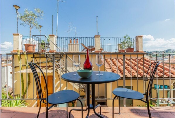 Un balcón con una mesa redonda y dos sillas, un jarrón rojo y vistas a edificios con tejados de terracota.