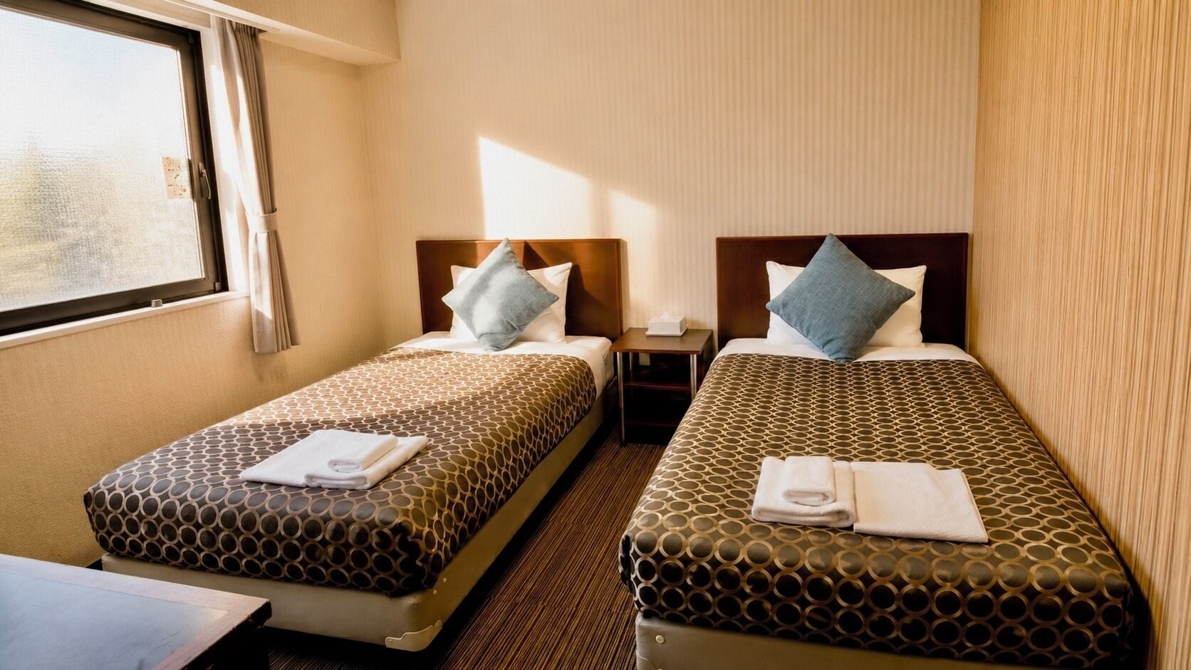 Habitación de hotel con dos camas, cada una con cubrecama y almohadas estampadas, una mesita de noche con lámpara y una ventana con cortinas.