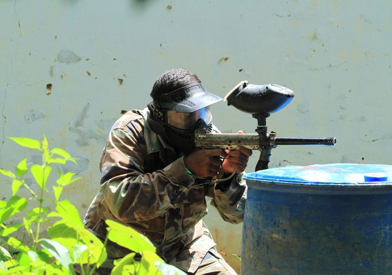 A person in camouflage paintball gear aiming a paintball gun at a blue barrel.
