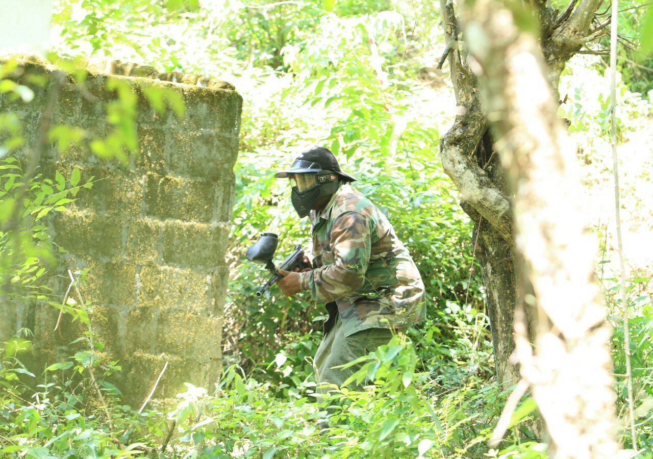 Una persona vestida con ropa de camuflaje y casco, agachada en un entorno natural exuberante y verde, posiblemente un campo de paintball o zona de entrenamiento táctico.
