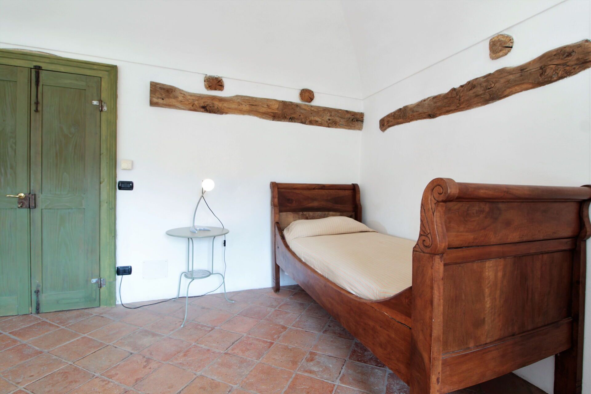 Une petite chambre avec un lit simple, une tête de lit en bois, une porte verte et une petite table avec une lampe.