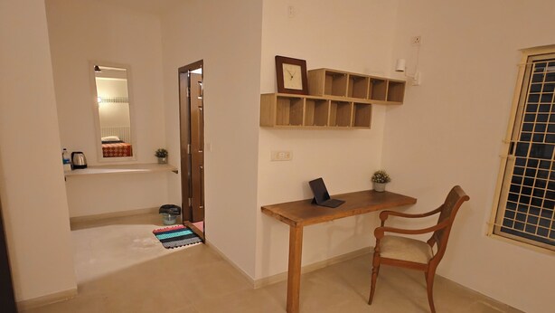A room with a wooden desk, a chair, a mirror, a shelf with a clock, and a window with a grid pattern. - Transit Stay (Bengaluru)