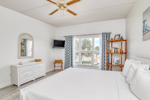 A bedroom with a large bed, a ceiling fan, a TV, a window with curtains, a bookshelf, and a dresser.