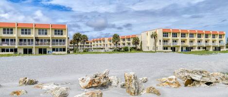A beachfront hotel with multiple-story buildings, a sandy foreground with scattered rocks, and a clear sky.
