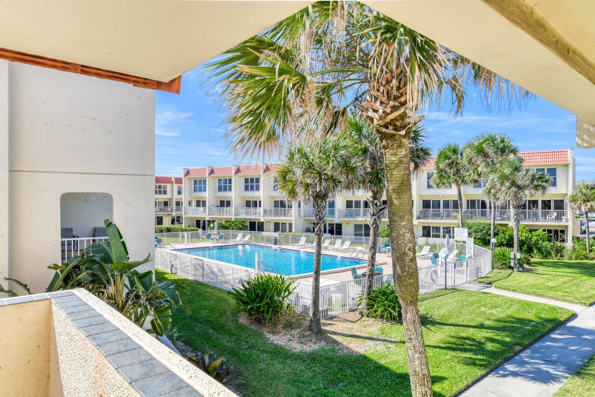 A pool area with palm trees, a clear blue sky, and a building with a red roof in the background.