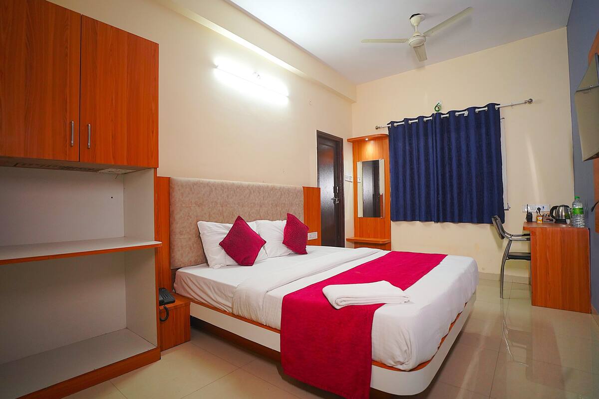 A hotel room with a bed, wooden wardrobe, a mirror, a desk with a kettle, and a ceiling fan.