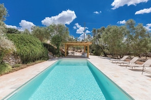 Une piscine rectangulaire avec une pergola en arrière-plan, entourée d’arbres et de chaises longues.