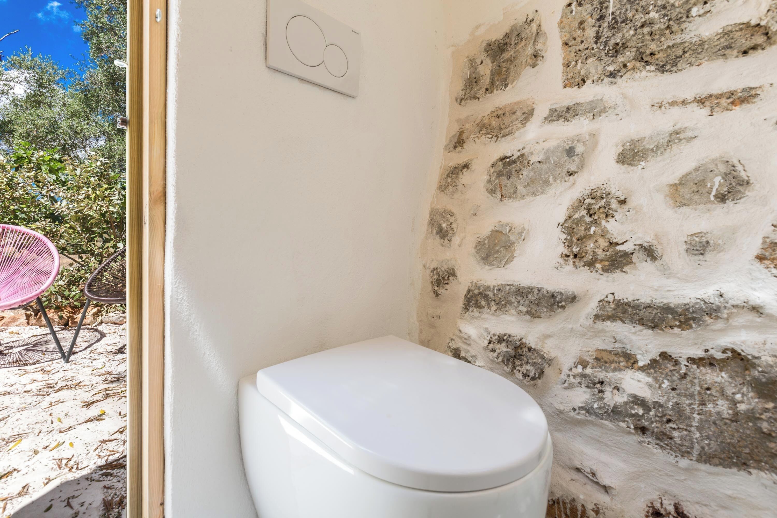 Un baño con inodoro blanco, pared de piedra y puerta que da a un espacio exterior con una silla y plantas.