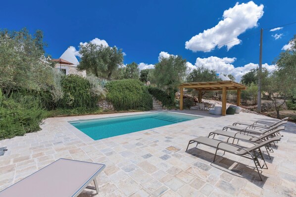 Piscina con sillas de descanso y una pérgola, rodeada de follaje y baldosas de piedra.