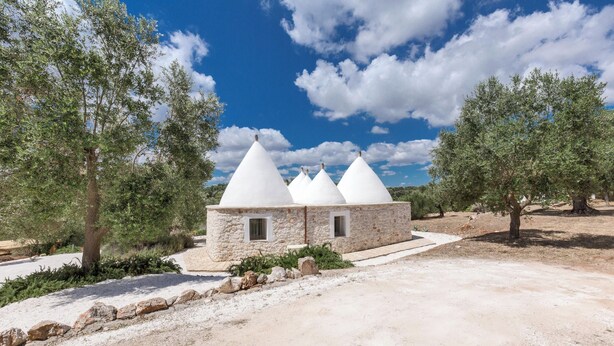 A stone building with three conical roofs, surrounded by olive trees and a clear sky.