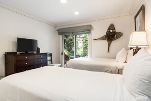 A hotel room with two beds, a TV on a dresser, a balcony with a view of greenery, and a decorative wall piece.