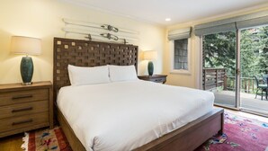 A bedroom with a large bed, wooden headboard, two bedside tables with lamps, a window with a view of greenery, and a balcony with outdoor furniture.