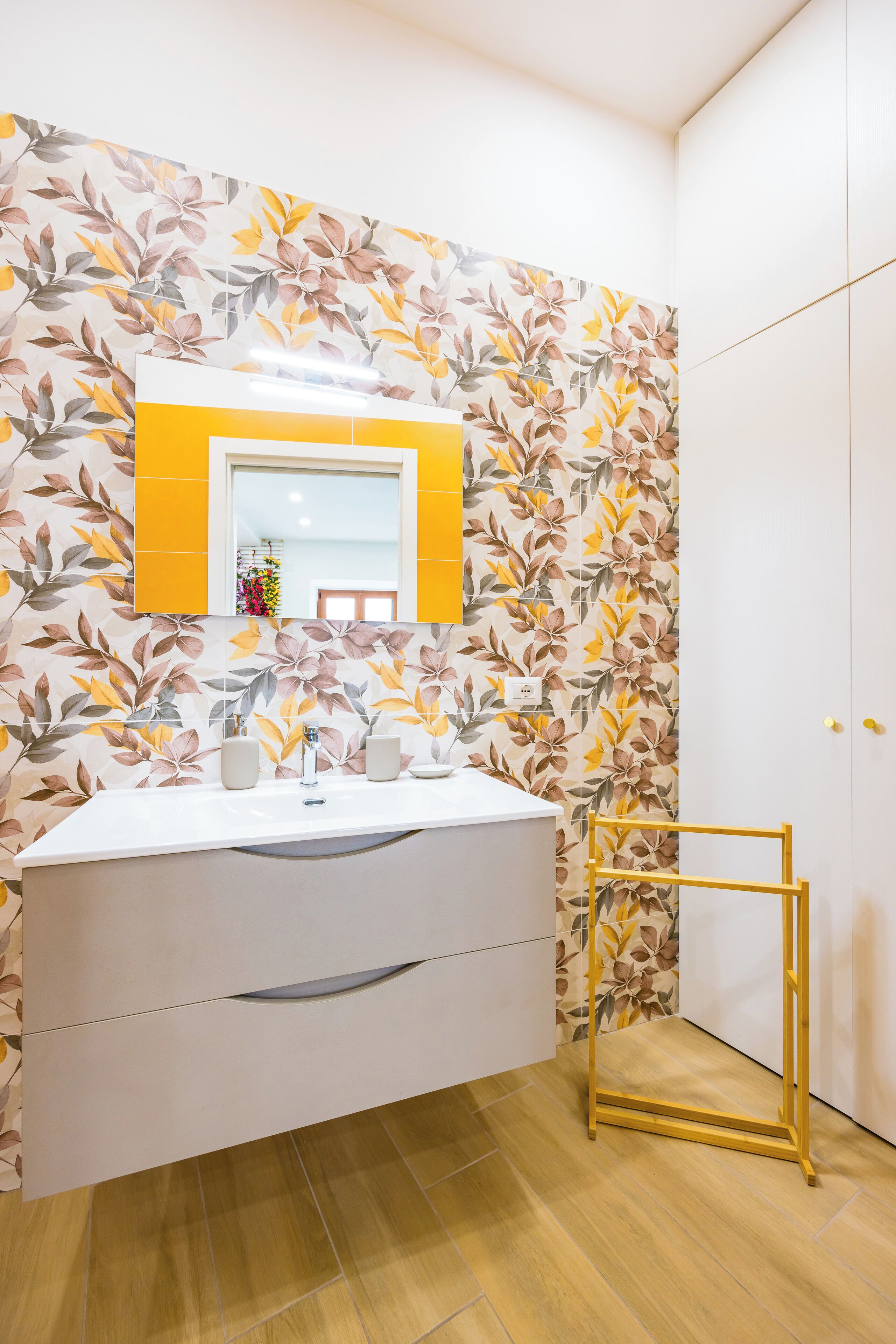 A bathroom with a floral wallpaper, a white vanity with a mirror, and a yellow towel rack.