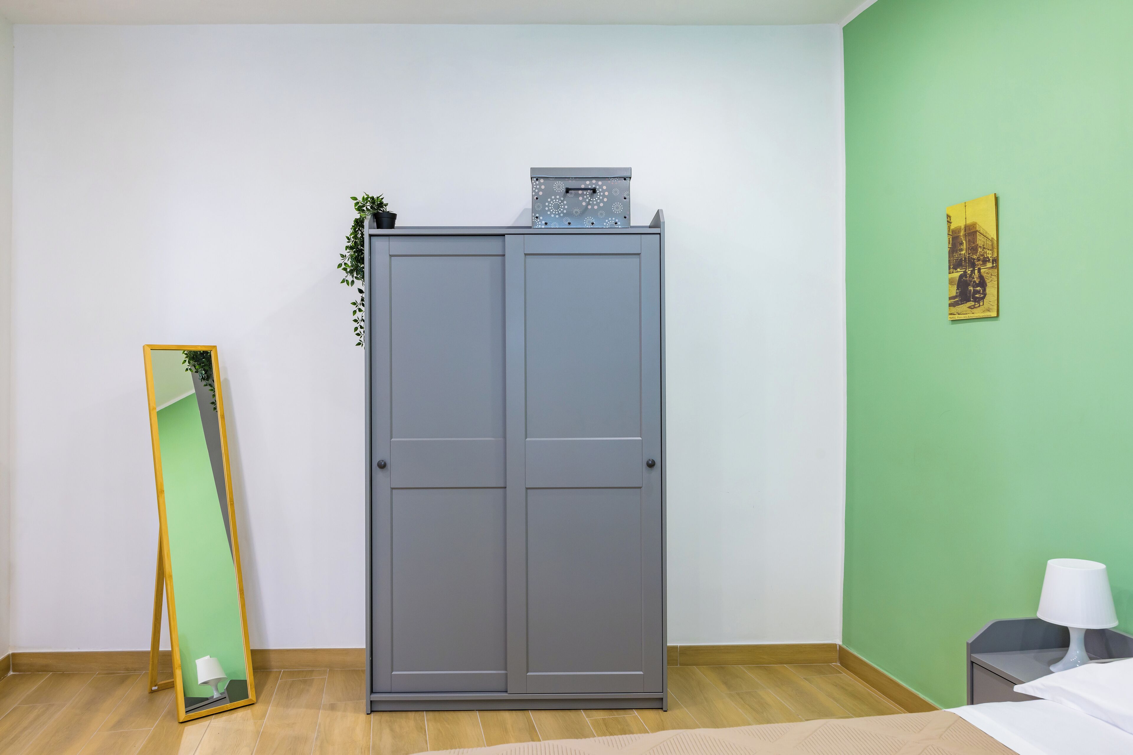 A room with a grey wardrobe, a green wall, a bed, and a nightstand with a lamp.