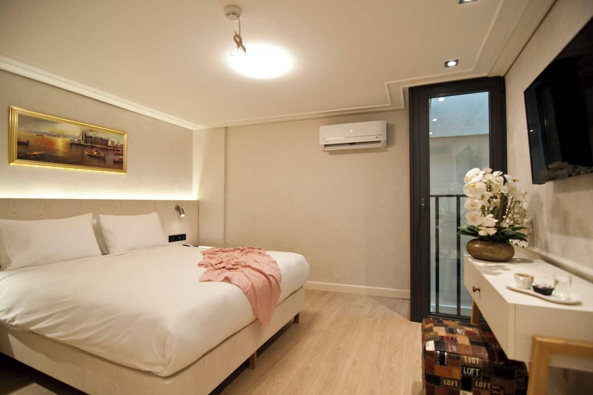 A hotel room with a bed, a bedside table with a vase of flowers, a wall-mounted air conditioner, and a painting above the bed.