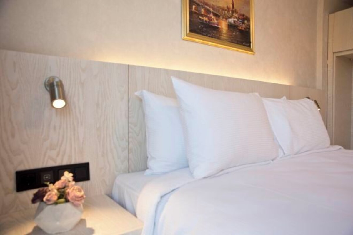A hotel room with a bed, white bedding, a nightstand with a vase of flowers, a wall-mounted light, and a framed picture above the bed.
