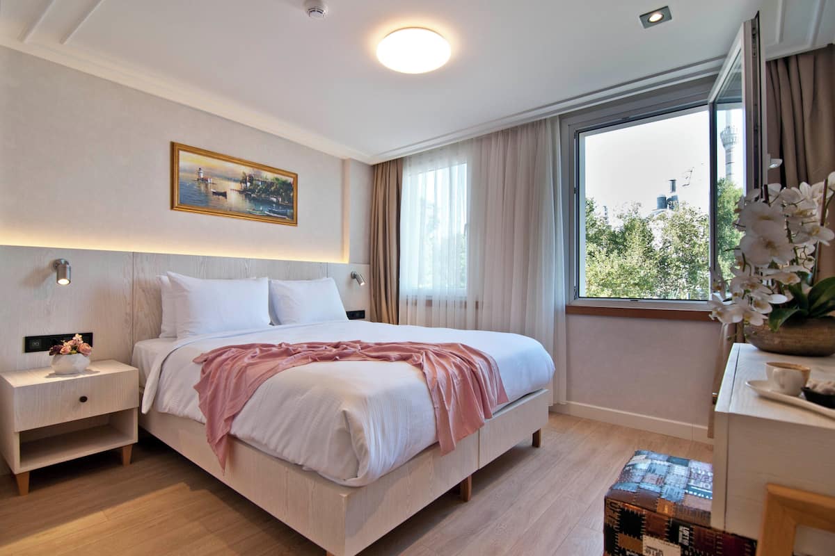 A modern hotel room with a large bed, a nightstand, a desk with a vase of flowers, and a window with a city view.