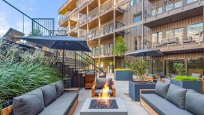 An outdoor seating area with modern furniture, a central fire pit, and a multi-story building in the background. - Luxury Penthouse w/ 2 Floors - Pool, Patio, Gym, Walk to Broadway (Nashville)