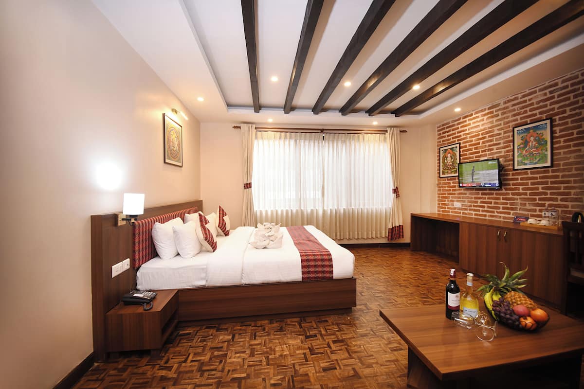 Habitación de hotel con piso de madera, una cama con ropa blanca, cabecera de madera, mesita de noche, una mesa con frutero, una botella de vino y televisión en la pared.
