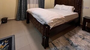 A bedroom with a bed, curtains, a nightstand, and a wall-mounted light. - Lux Suites Kipande Family Apartments (Nairobi)