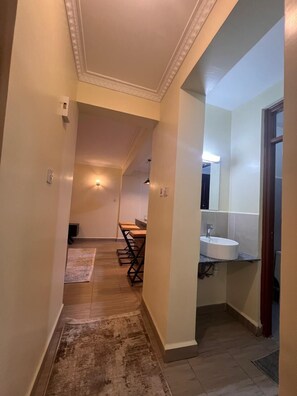 A hallway with a wooden floor, a dining area with tables and chairs, and a bathroom with a sink and mirror. - Lux Suites Kipande Family Apartments (Nairobi)