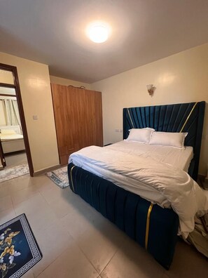 A bedroom with a bed, a wooden wardrobe, a nightstand, and a wall-mounted light fixture. - Lux Suites Kipande Family Apartments (Nairobi)