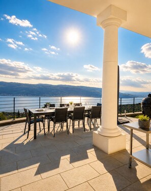 Terraço com mesa posta para refeições, com vista para um corpo d'água e colinas.