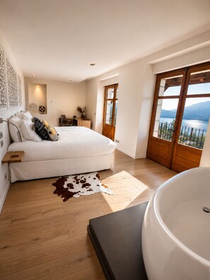 Quarto com uma cama grande, piso de madeira, tapete de couro de vaca, banheira e vista para um lago através de uma janela ampla.