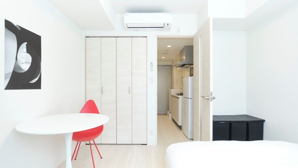 A modern hotel room with a bed, a small table with a red chair, a wardrobe, and a kitchenette.