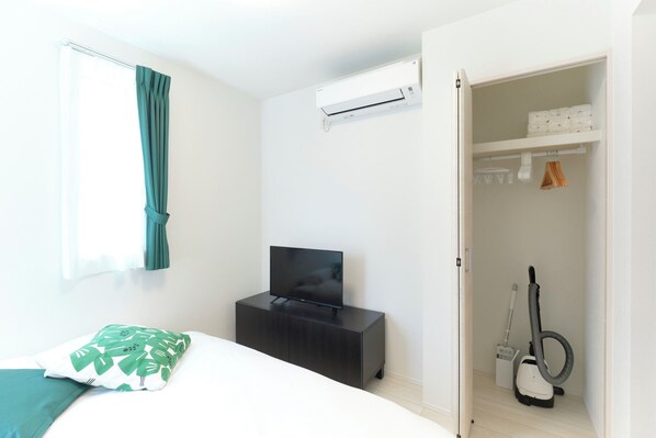 A bedroom with a bed, a TV on a cabinet, an air conditioner, and a closet with a vacuum cleaner.
