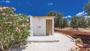 Ein kleines, weißes Gebäude mit einem Zimmer, einer Holztür und einem kleinen Fenster, umgeben von einem Kiesweg und Kakteen.