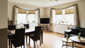 Una sala de estar con comedor, un sofá de cuero negro, un televisor sobre un mueble bajo y ventanas grandes con cortinas.