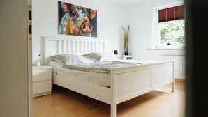 Una cama blanca con cabecera, una mesita de noche con un florero, y una ventana con persianas.