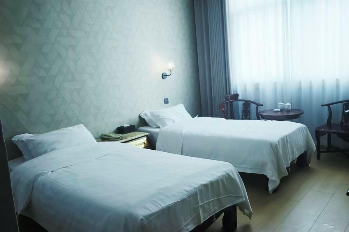 A hotel room with two beds, a small table, a chair, and a window with curtains.