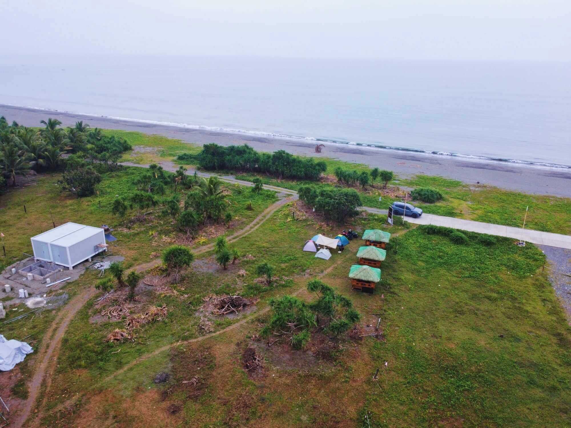 An aerial view of a coastal area with a large white building, several small huts with thatched roofs, and a paved road leading to the beach.