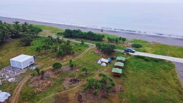 An aerial view of a coastal area with a large white building, several small huts with thatched roofs, and a paved road leading to the beach.