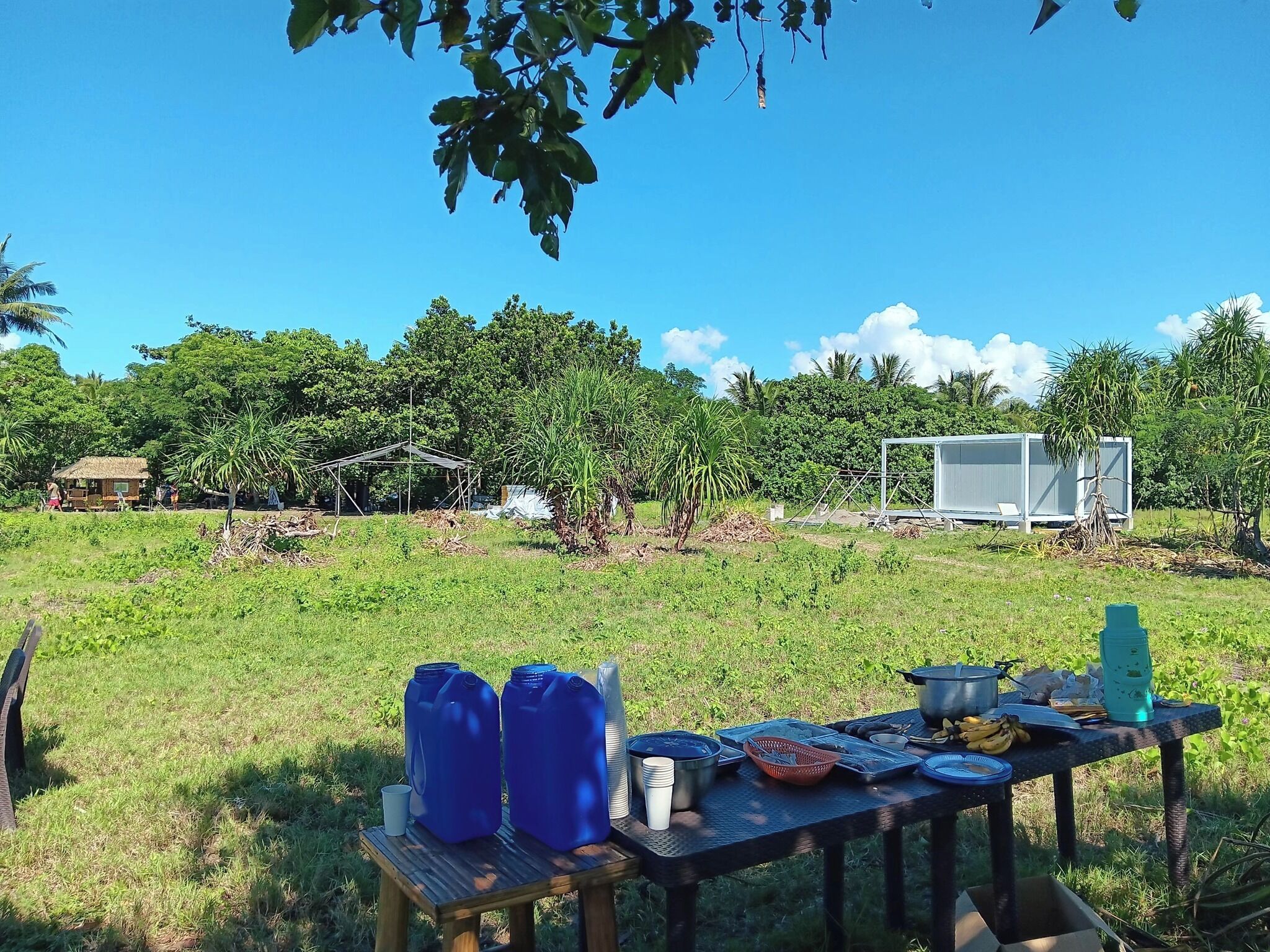 A wooden table with blue containers, kitchenware, and a water bottle in an outdoor setting with trees and a clear sky.