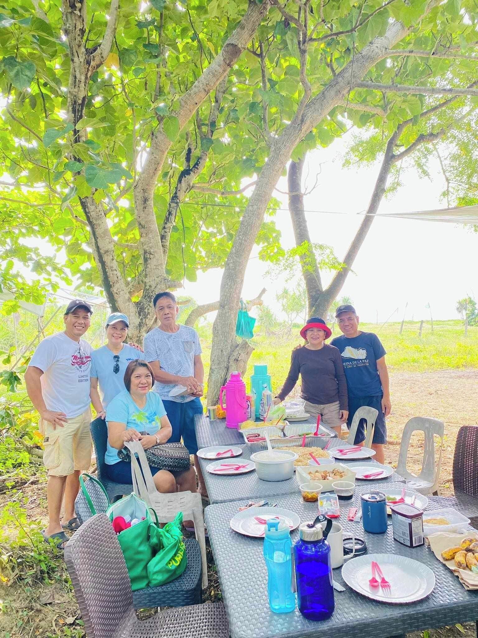 Un gruppo di persone riunito attorno a un tavolo imbandito di cibo e bevande, all'aperto sotto gli alberi.